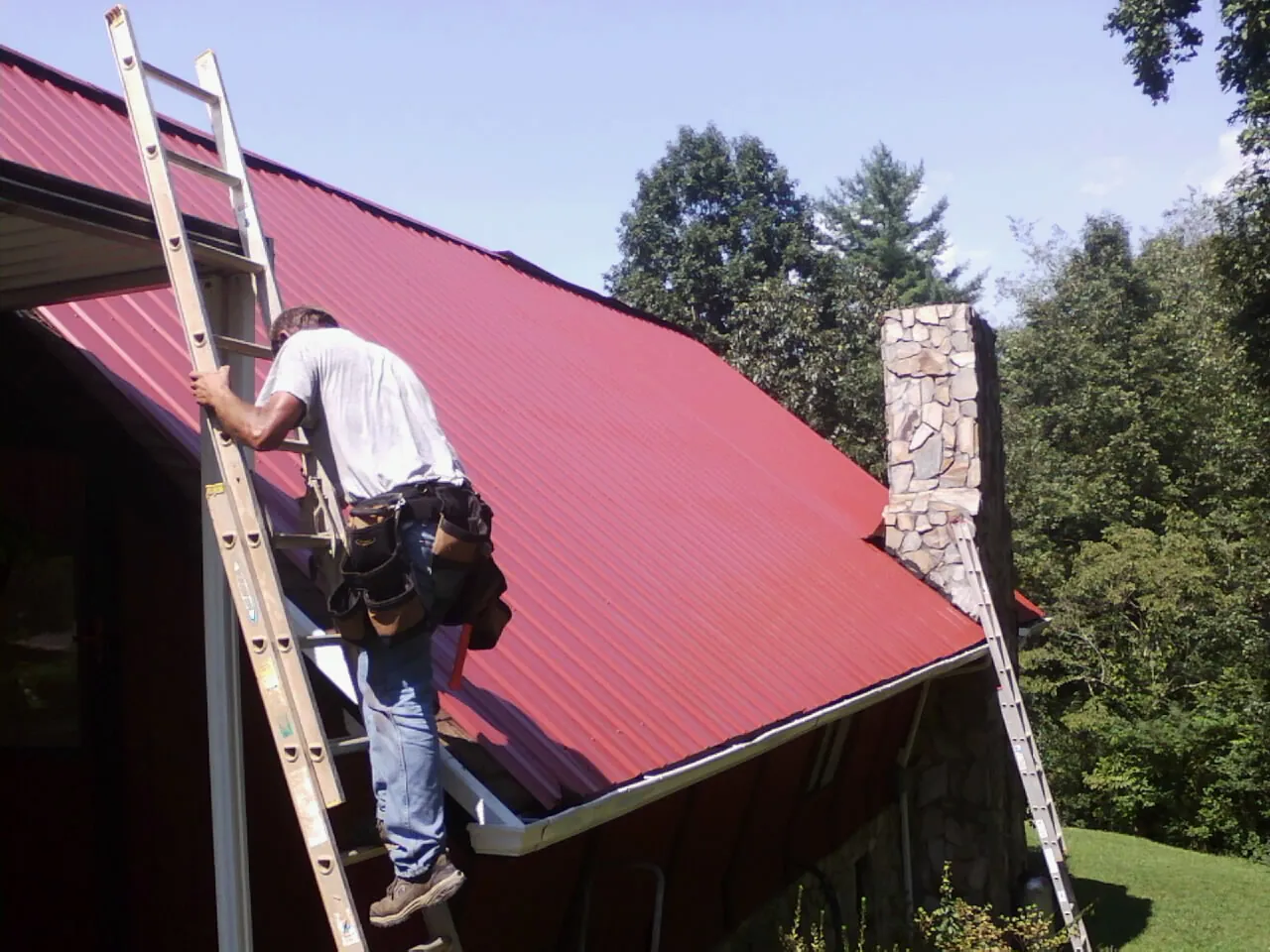 Roofer on a ladder beside a red metal roof and stone chimney — Soffit & Fascia Repair by Roofing Brothers in Danvers